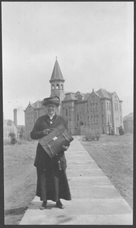 When Grace Raymond Hebard arrived at the University of Wyoming in 1891, Old Main, shown behind her, was the university. Photo File – Hebard, Grace Raymond, University of Wyoming, American Heritage Center. Woman in glasses, long black dress, jacket, hat and gloves posesWhen Grace Raymond Hebard arrived at the University of Wyoming in 1891, Old Main, shown behind her, was the university. Photo File – Hebard, Grace Raymond, University of Wyoming, American Heritage Center. holding a leather briefcase, with an ornate stone building in the background